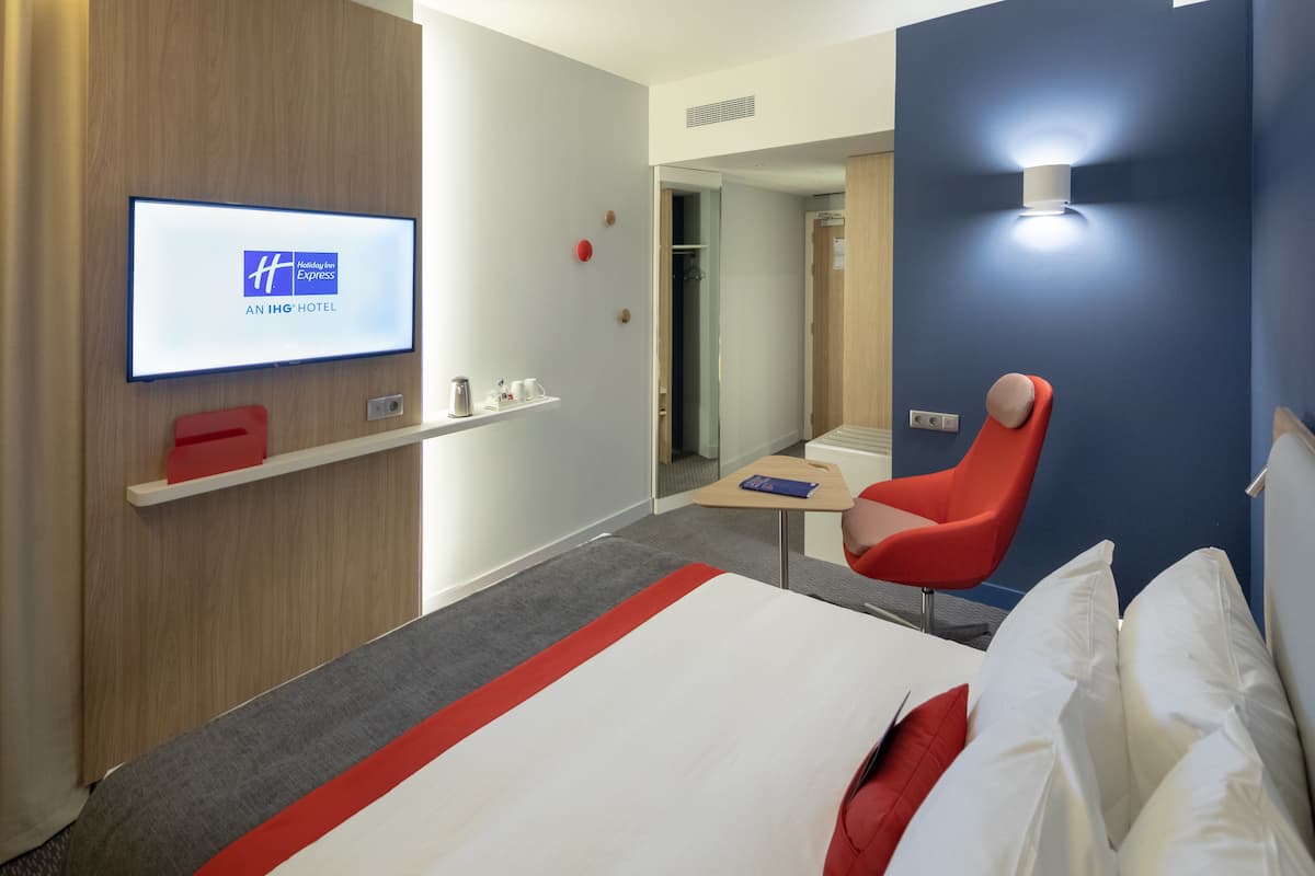 Una habitación de hotel moderna con una cama, una silla roja, un televisor mostrando el nombre del hotel y una pequeña mesa con una cafetera.