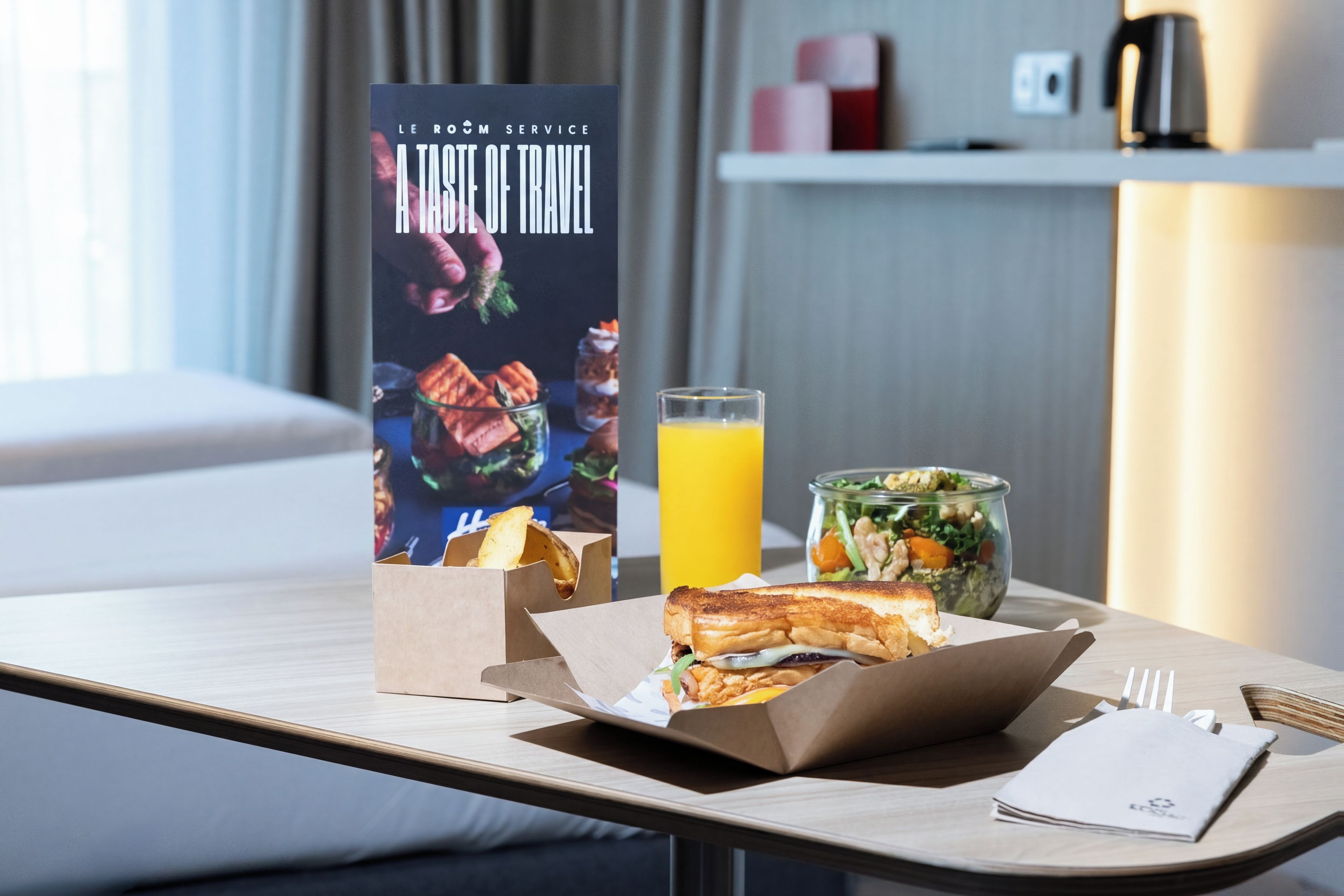 Habitación de hotel con cama, una mesa y opciones de desayuno, incluyendo un sándwich, un vaso de zumo de naranja y una ensalada.