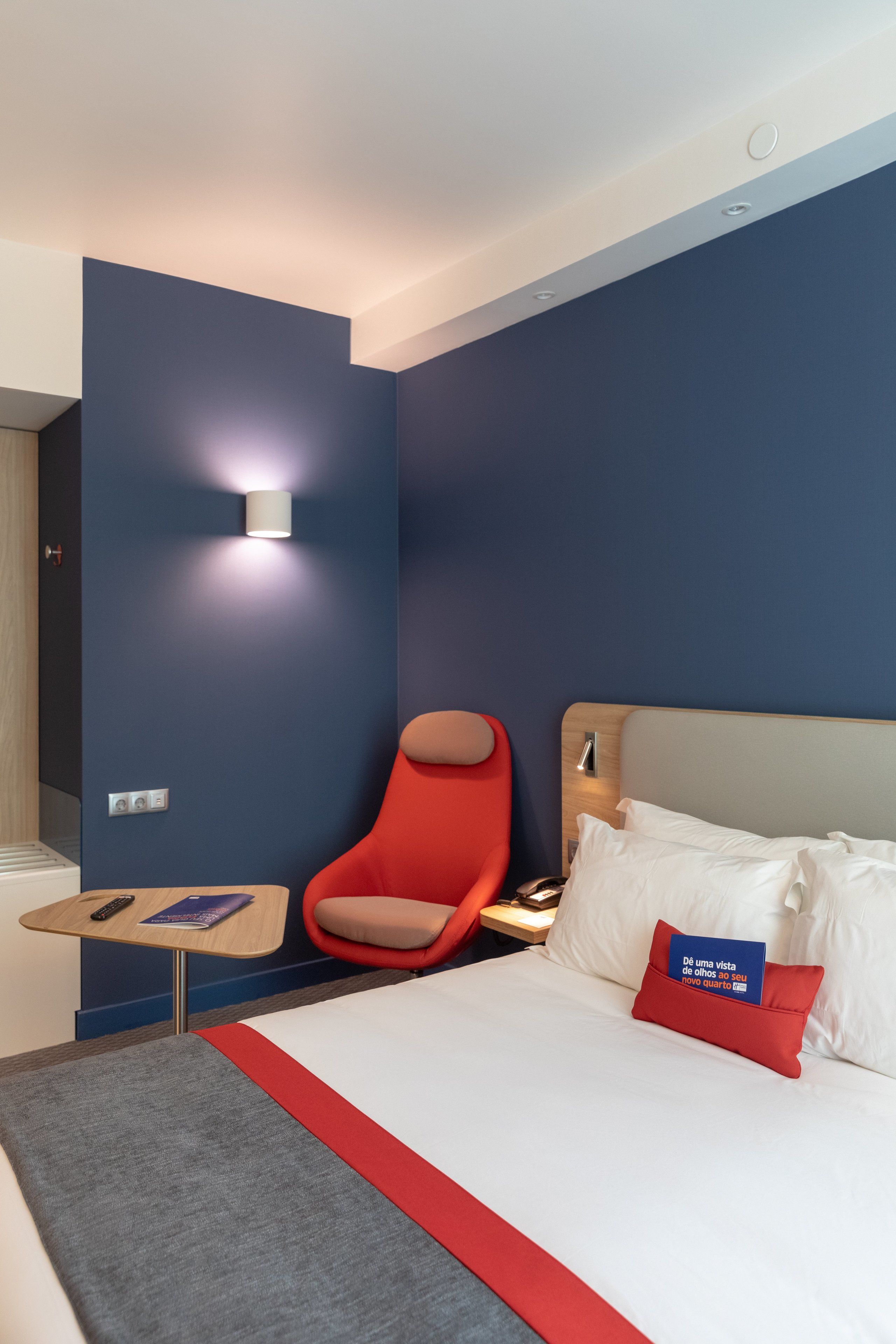 A hotel room with a bed, a red chair, a small table, and a wall-mounted light.