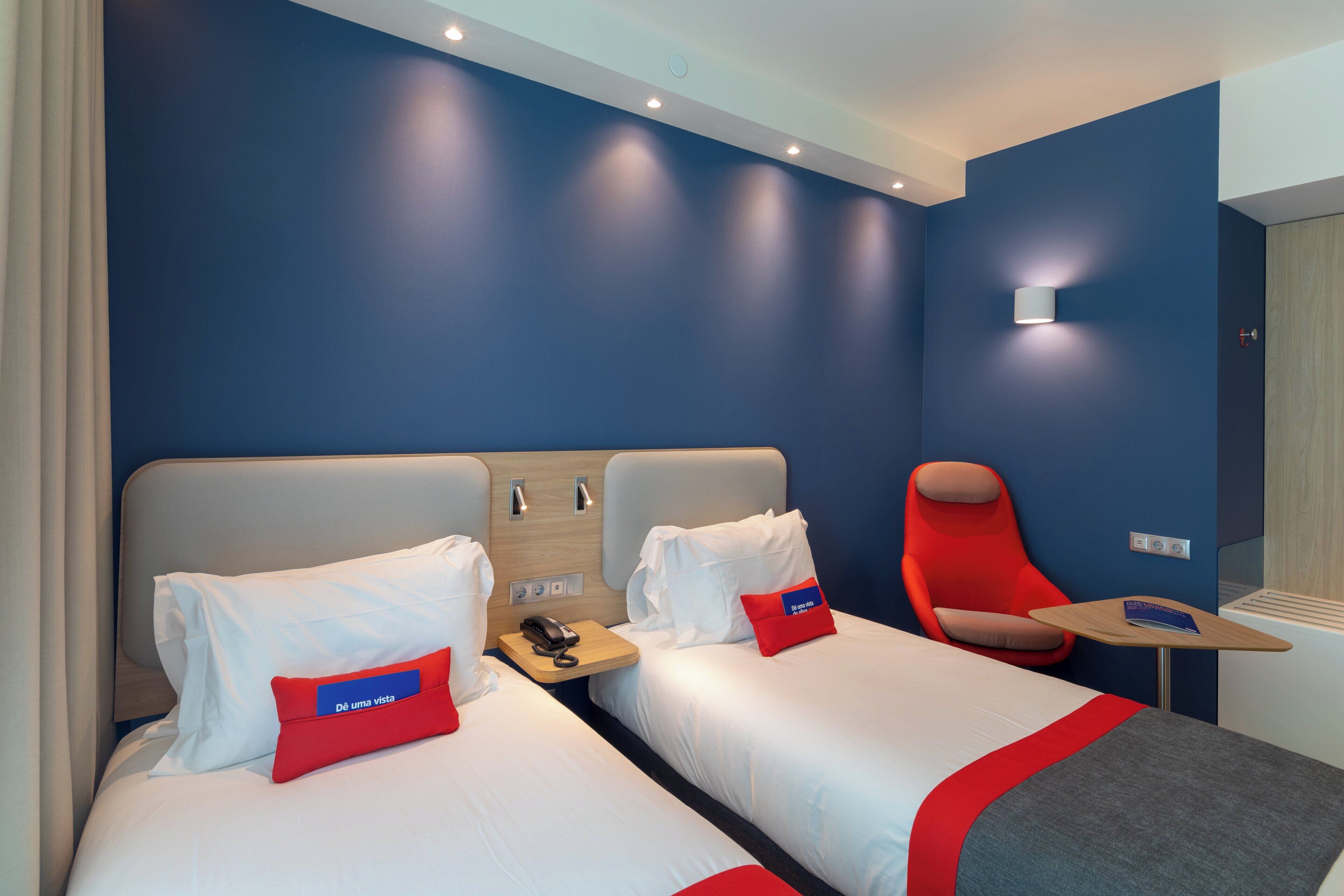 A hotel room with two beds, a red chair, a small table, and a wall-mounted phone.