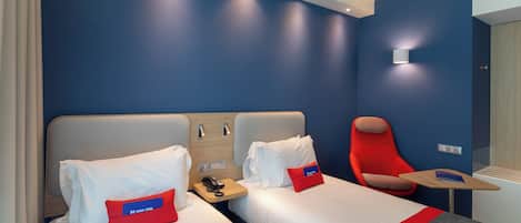 A hotel room with two beds, a red chair, a small table, and a wall-mounted phone.