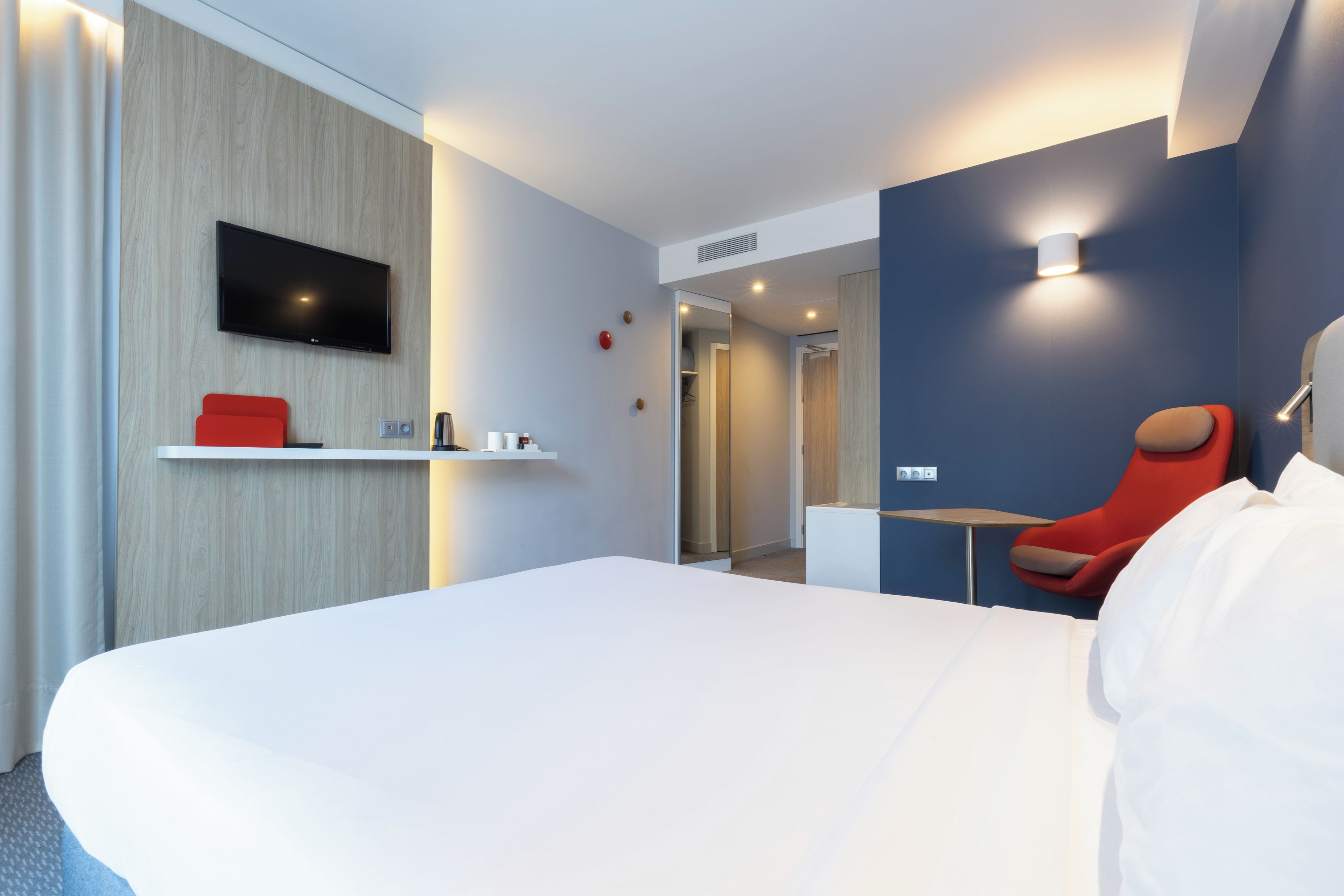 Habitación de hotel moderna con una cama grande, un televisor de pantalla plana montado en la pared, una silla roja y una pequeña mesa.