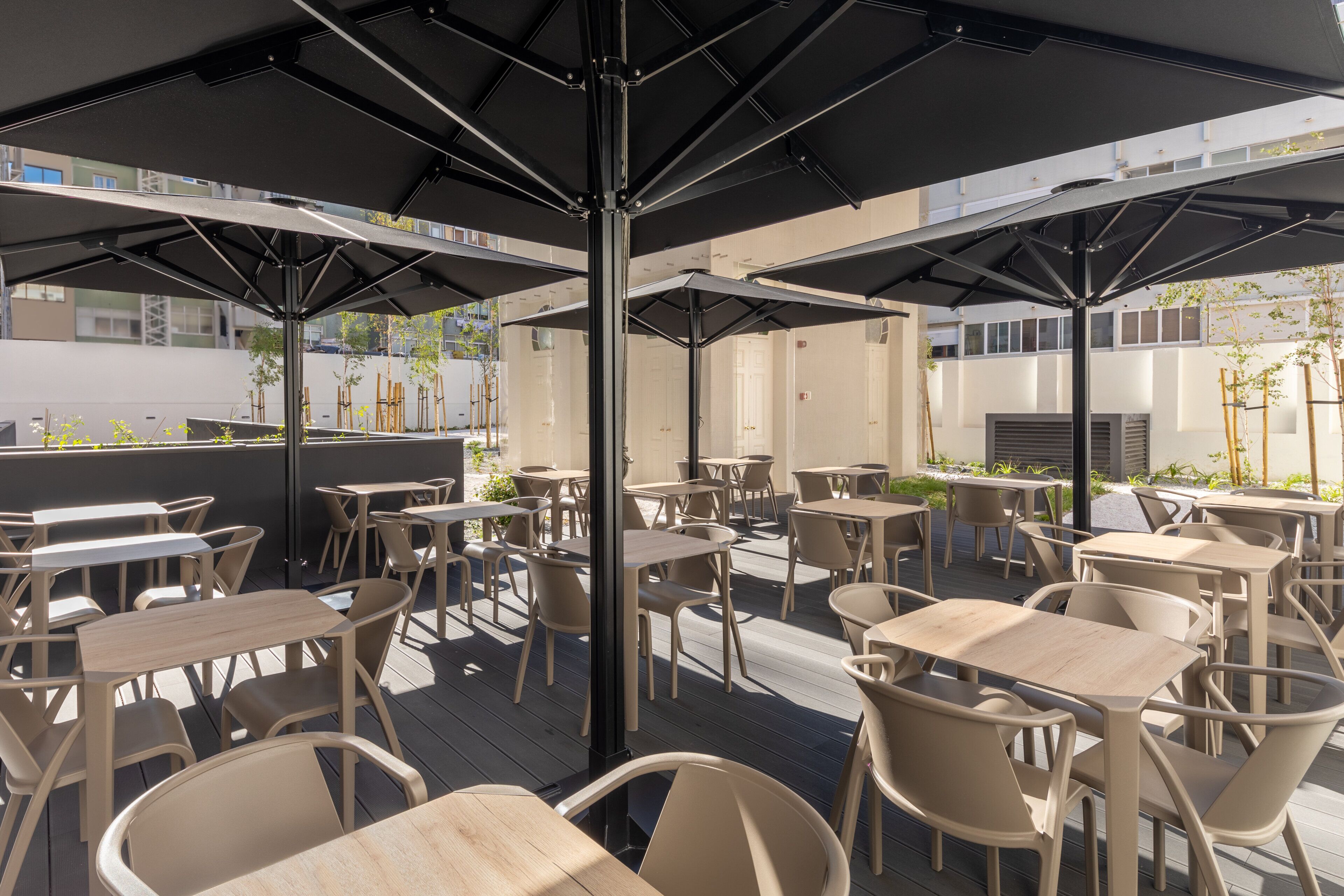 An outdoor seating area with multiple tables and chairs, shaded by umbrellas, and a view of buildings in the background.