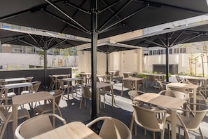 Un espace extérieur aménagé avec plusieurs tables et chaises, protégé du soleil par des parasols, et offrant une vue sur les bâtiments en arrière-plan.