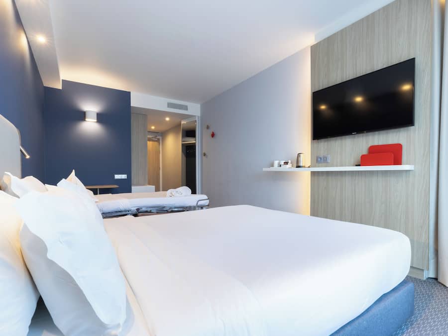 Une chambre d’hôtel avec un grand lit, un téléviseur à écran plat fixé au mur et une petite étagère sur laquelle se trouve un objet rouge.