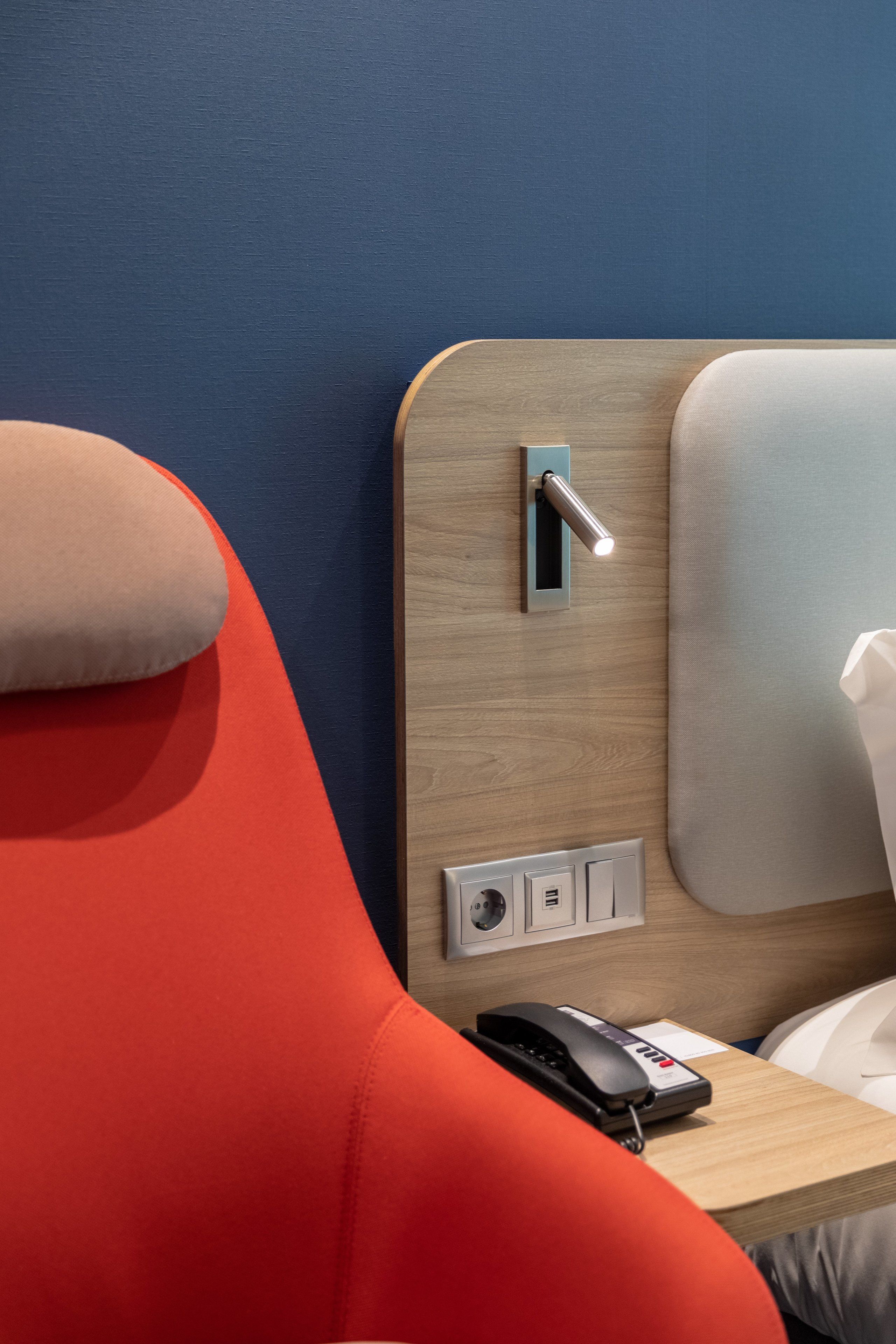 A modern hotel room with a wooden headboard, a red chair, a bedside table with a phone and a power outlet.