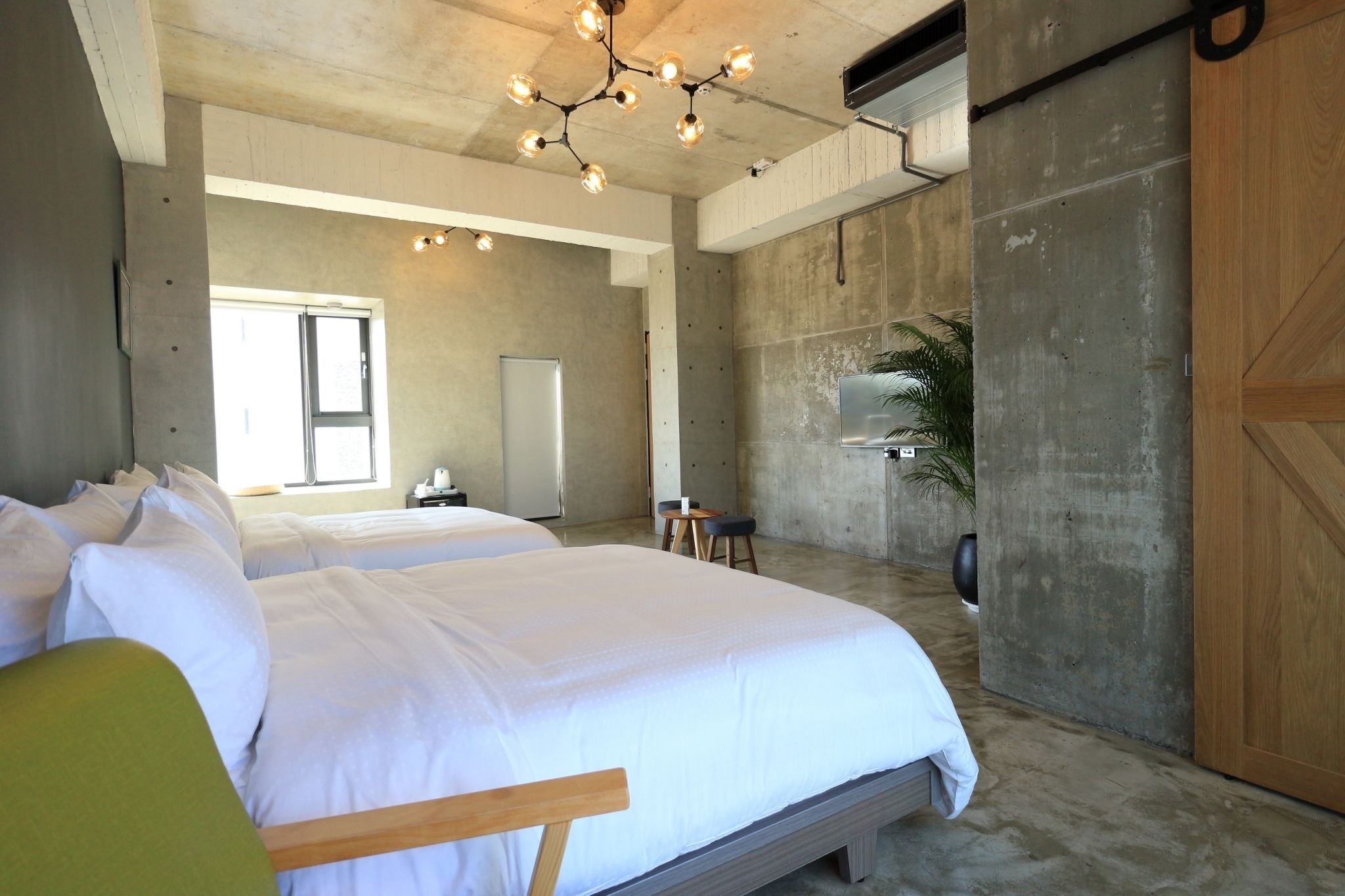 A modern hotel room with two beds, a concrete accent wall, a wooden door, and a ceiling light fixture.