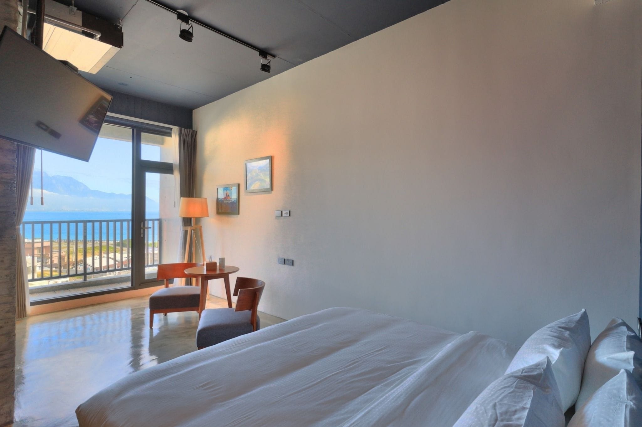 A hotel room with a large bed, a desk, a chair, a lamp, and a view of the sea through a balcony.