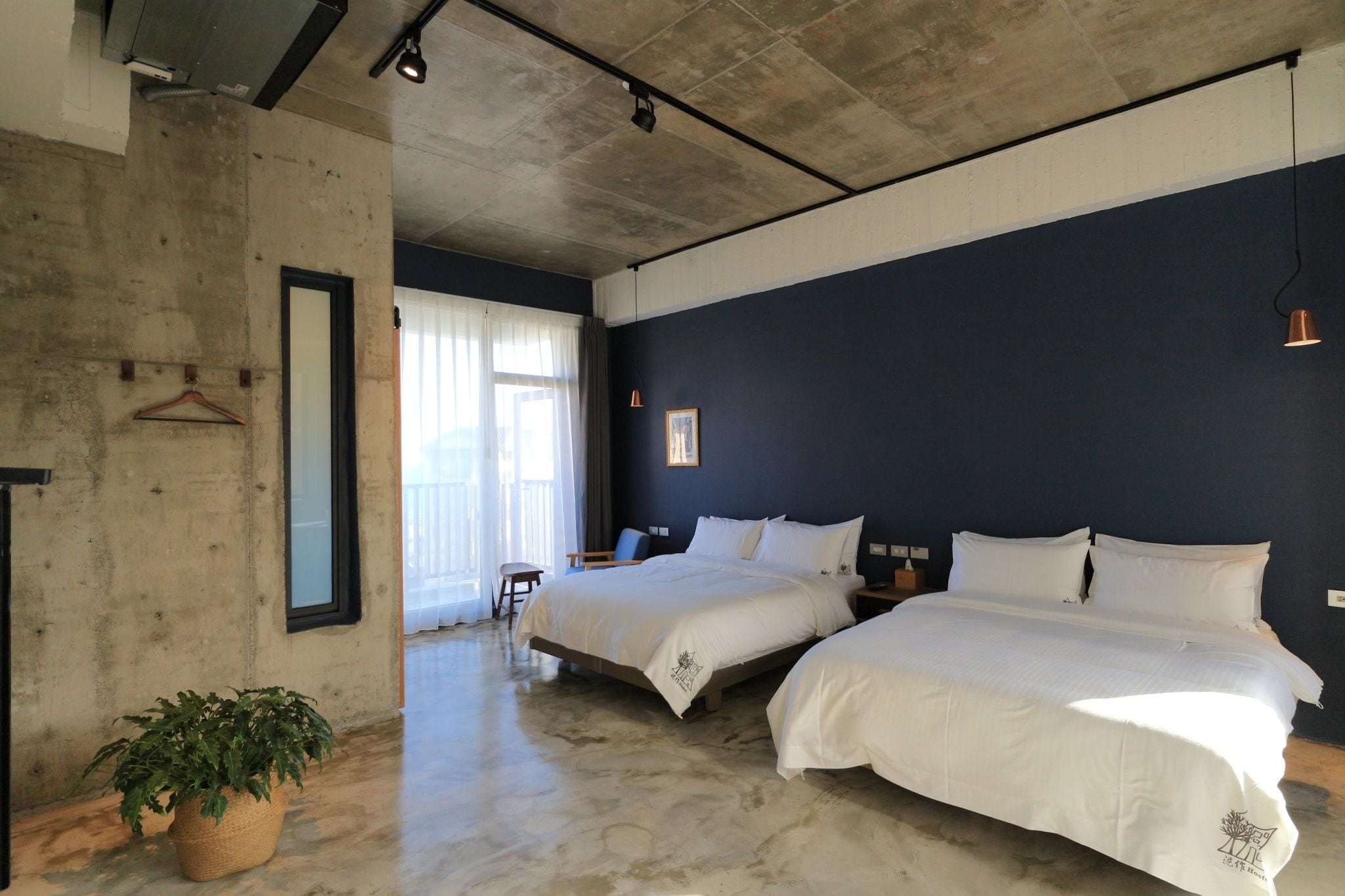 Habitación de hotel con dos camas, una planta en maceta y un ventanal con cortinas.