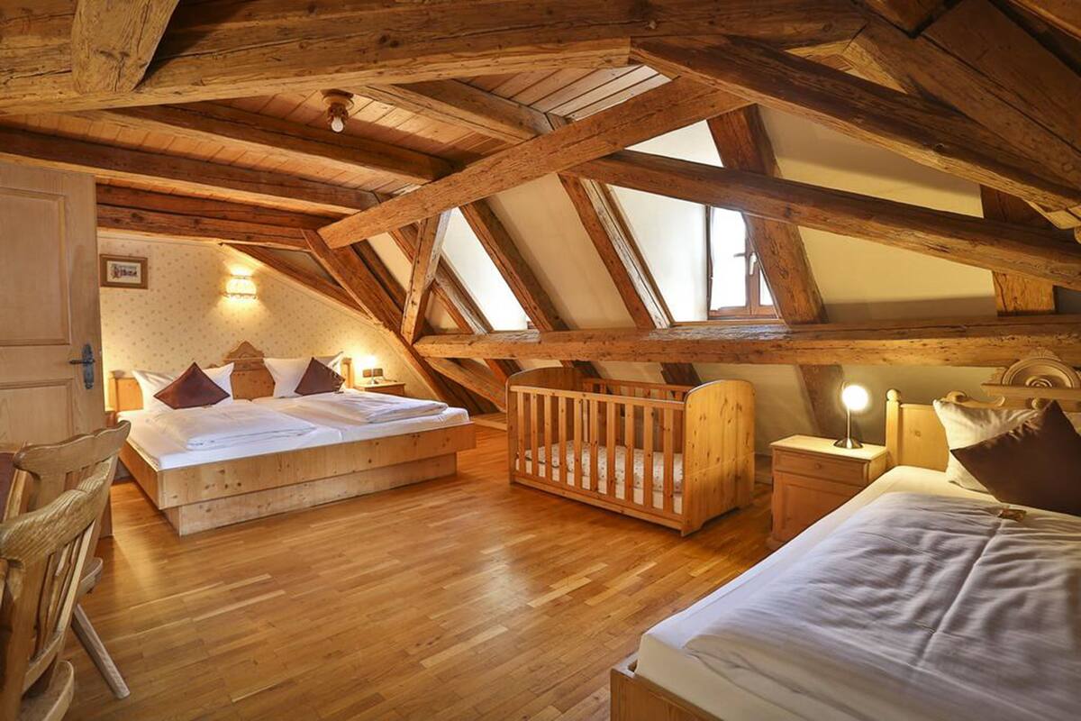 Una habitación con techo alto, vigas de madera, una cama grande, una cuna y un escritorio con lámpara.