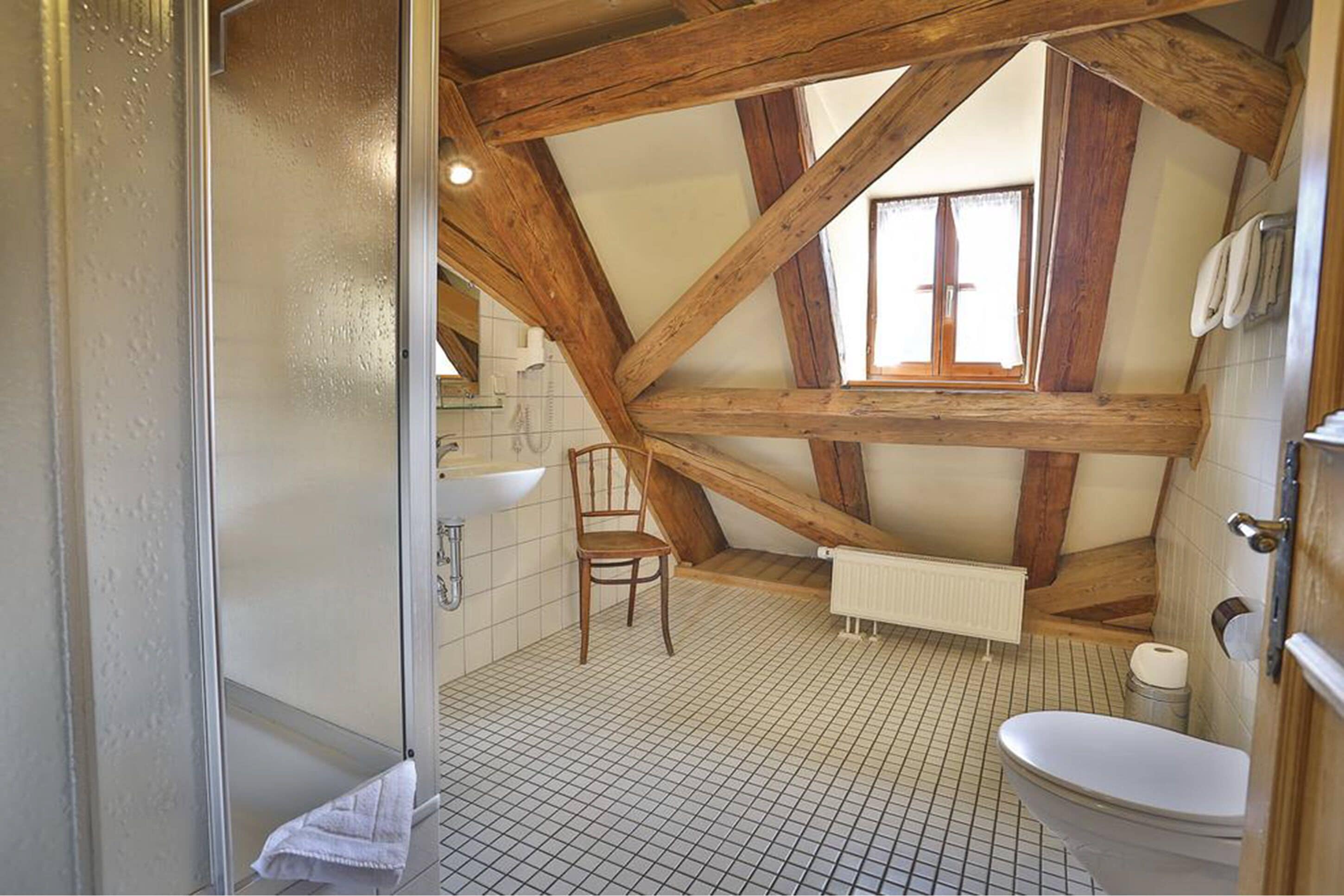 Un baño con techo inclinado, una silla de madera, un inodoro y una zona de ducha.