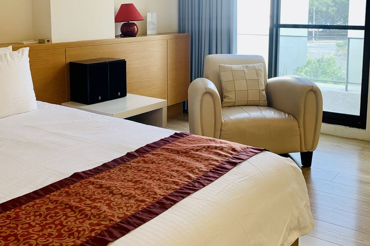 Una camera d'albergo con un letto grande, una scrivania con una lampada, una sedia e vista all'esterno attraverso una grande finestra.
