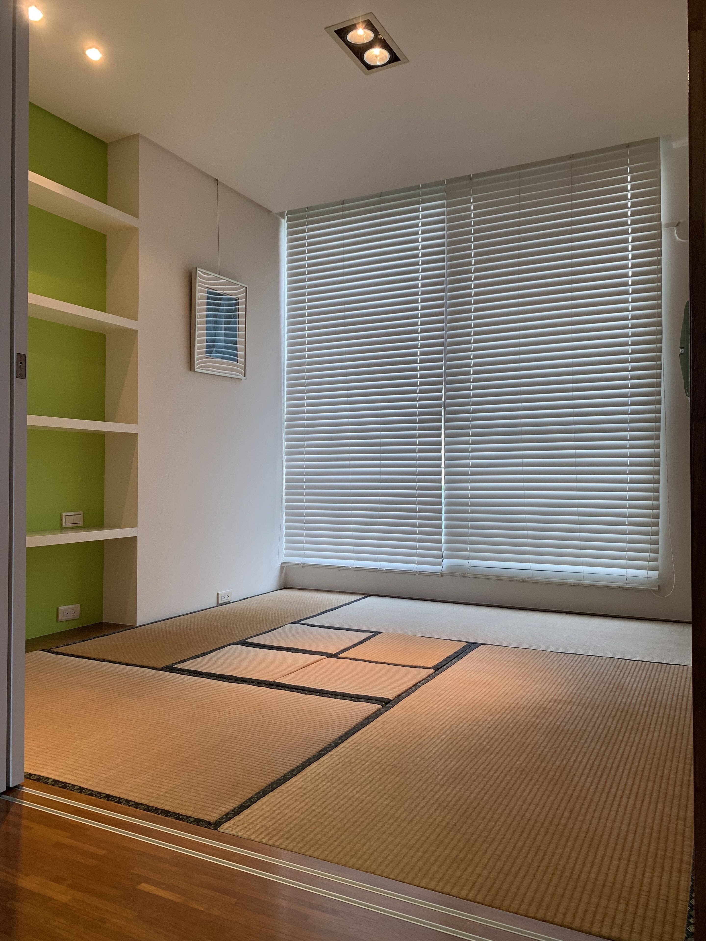 A room with tatami mats, a large window with blinds, green shelves, and recessed ceiling lighting.