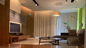 Una habitación de hotel moderna con televisión de pantalla plana, una mesa consola de madera, una mesa redonda de centro, un sofá y un ventanal con cortinas.
