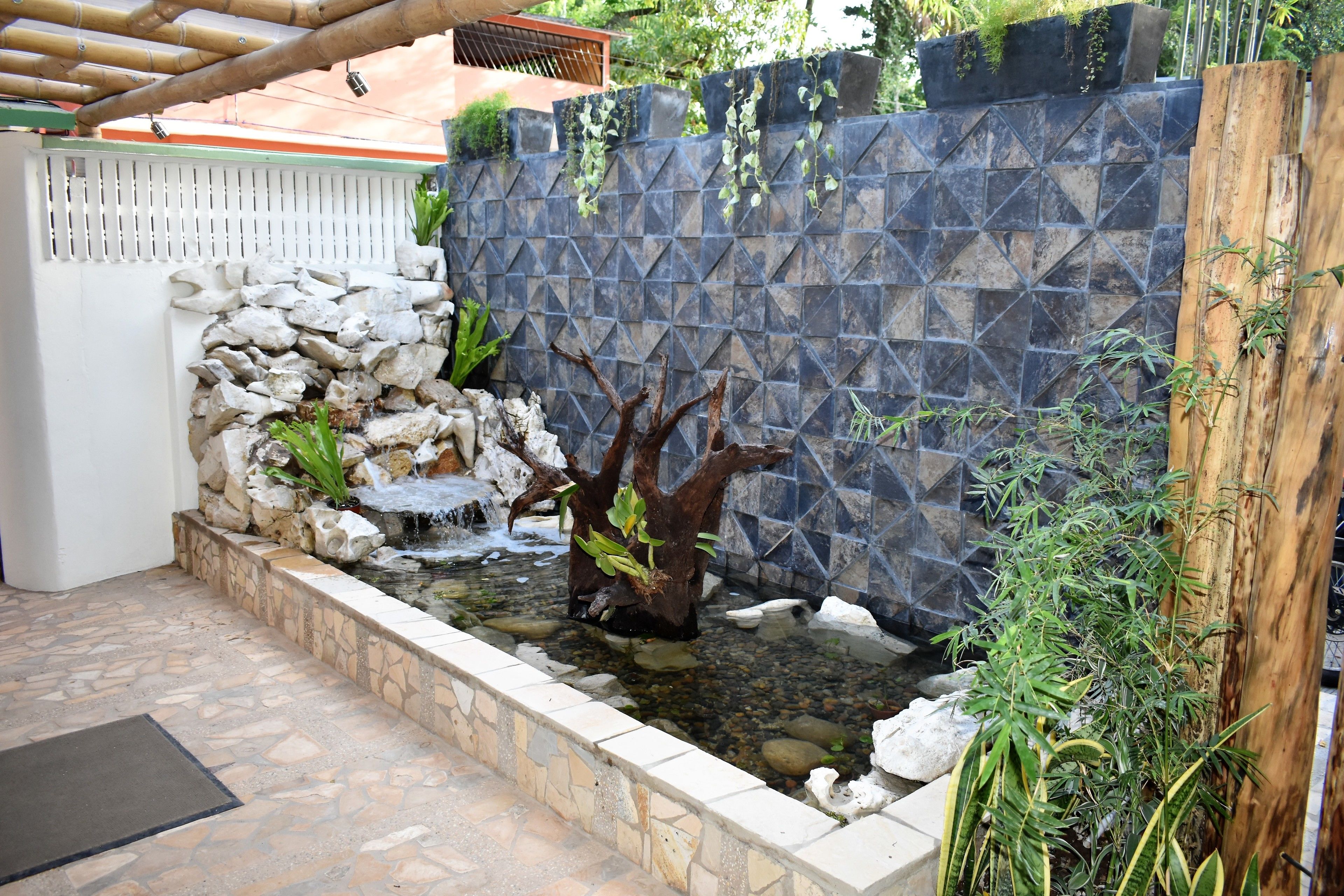 A stone-tiled patio with a small water feature, a wooden fence, and various plants.