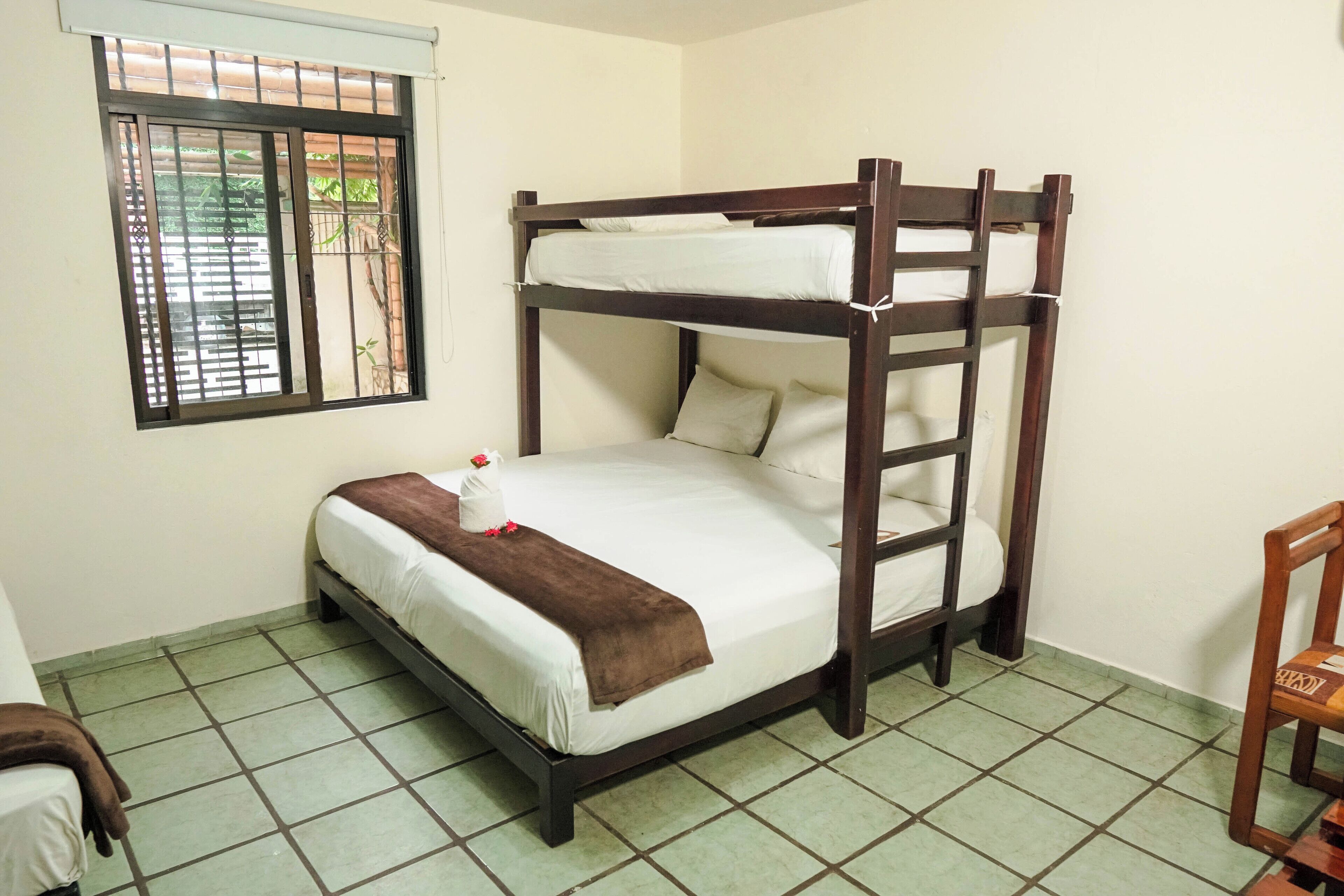 Uma cama beliche com janela, uma cadeira e piso ladrilhado.
