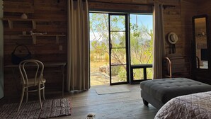 A rustic room with wooden walls, a large sliding glass door leading to an outdoor area, a bed, a chair, and a small table with a vase. - Rancho Cien Piedras (Valle de Guadalupe)