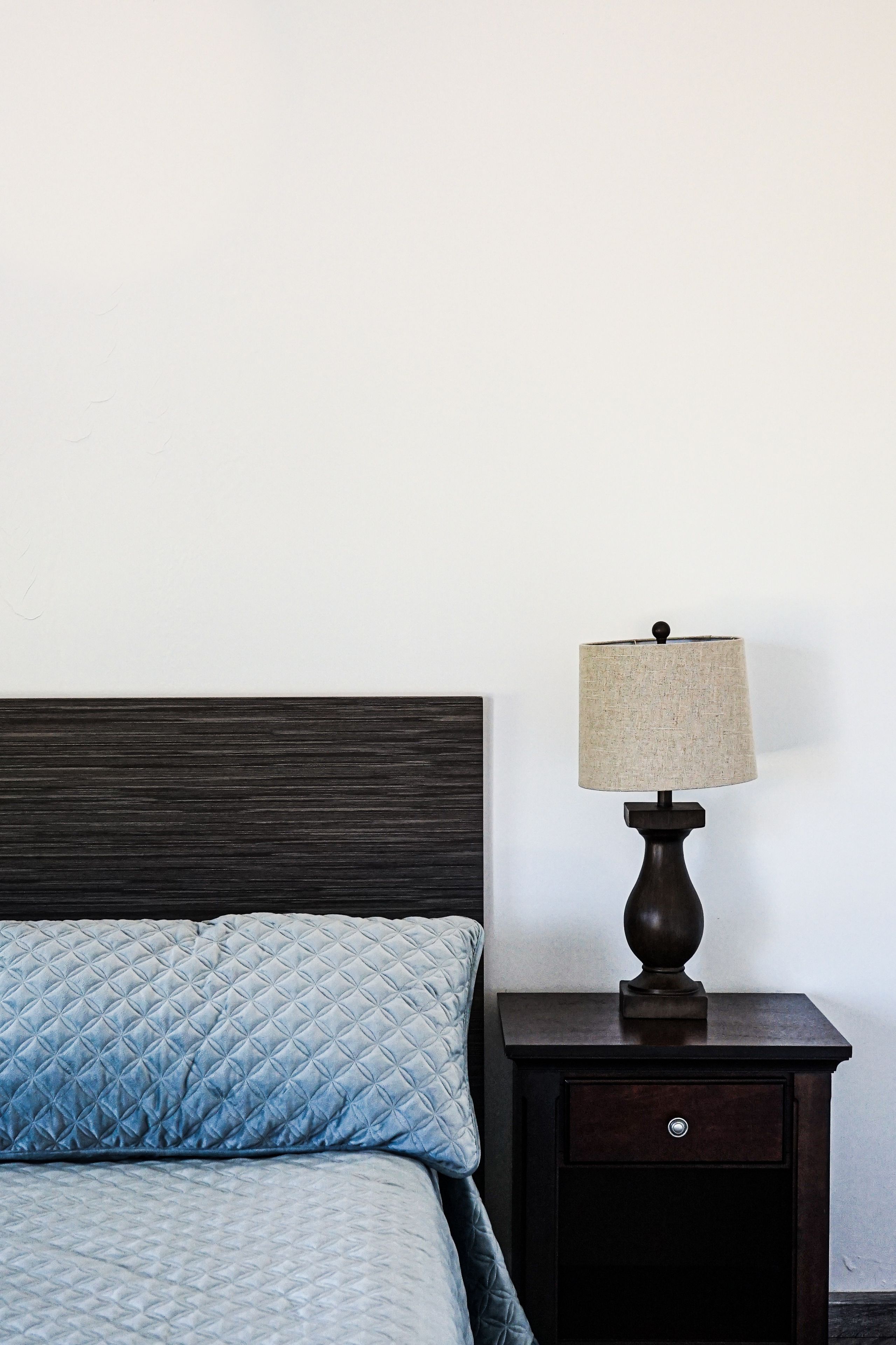 A bed with a quilted blue cover, a wooden nightstand with a lamp, and a dark wooden headboard.