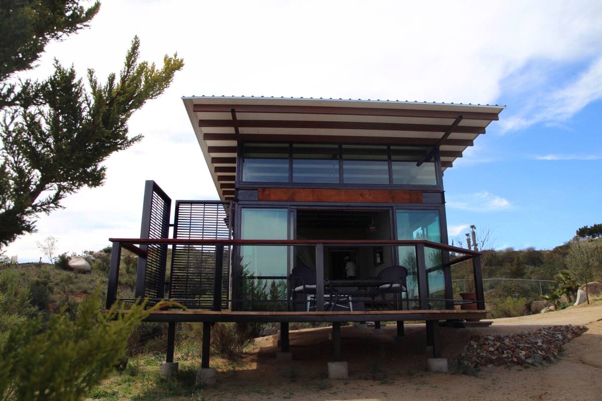 A modern, two-story house with a glass facade, a deck with outdoor seating, and a surrounding natural landscape.