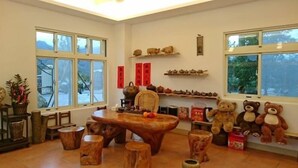 Una habitación acogedora con una mesa de madera, sillas y un gran oso de peluche. Hay ventanas, un estante con adornos y una planta en maceta.