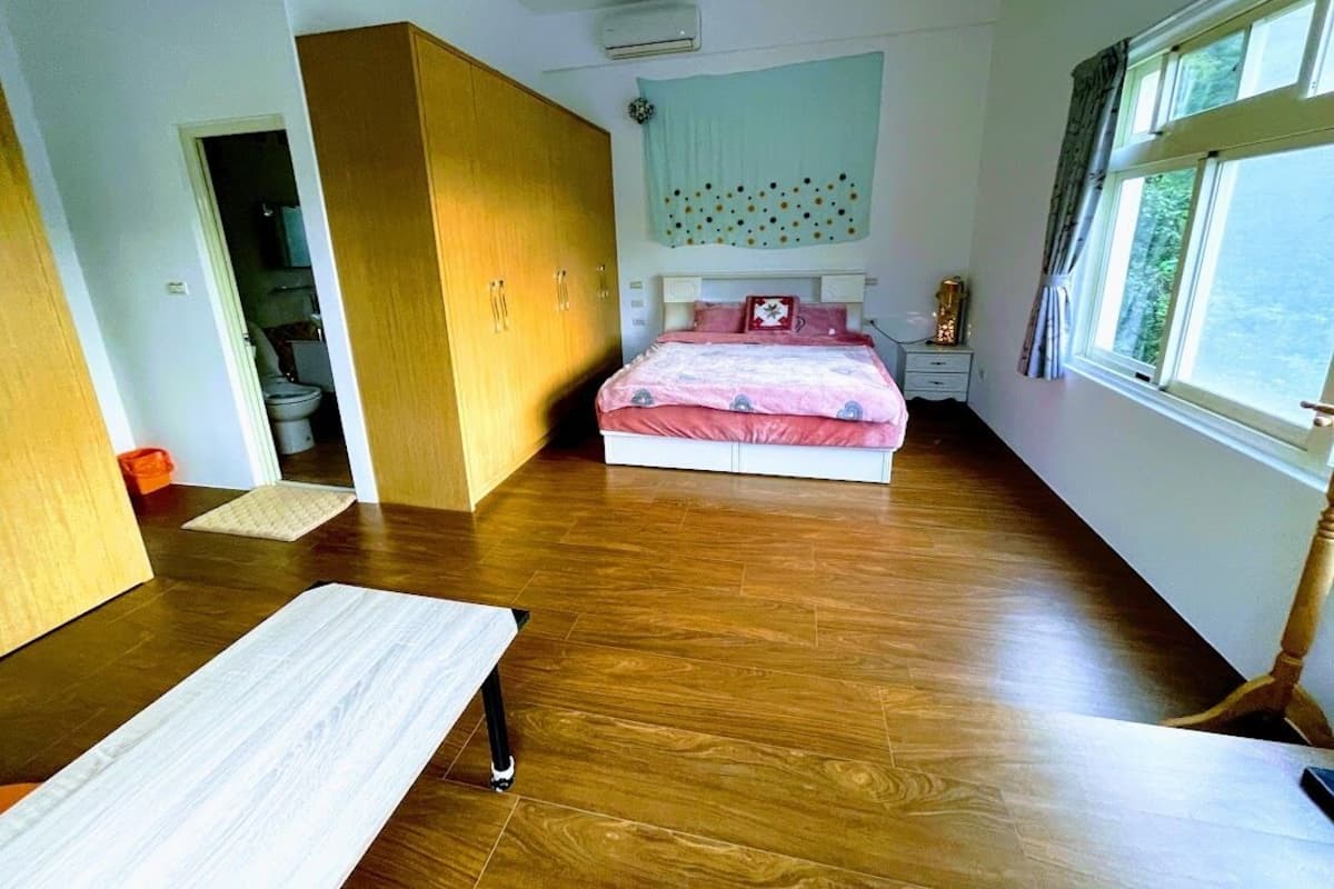A bedroom with a wooden floor, a bed with pink bedding, a white bedside table, a nightstand, and a window with curtains.