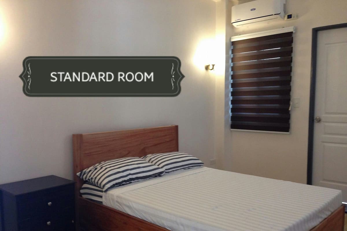 A standard room with a wooden bed, striped pillows, a dark nightstand, a wall-mounted air conditioner, and a window with blinds.