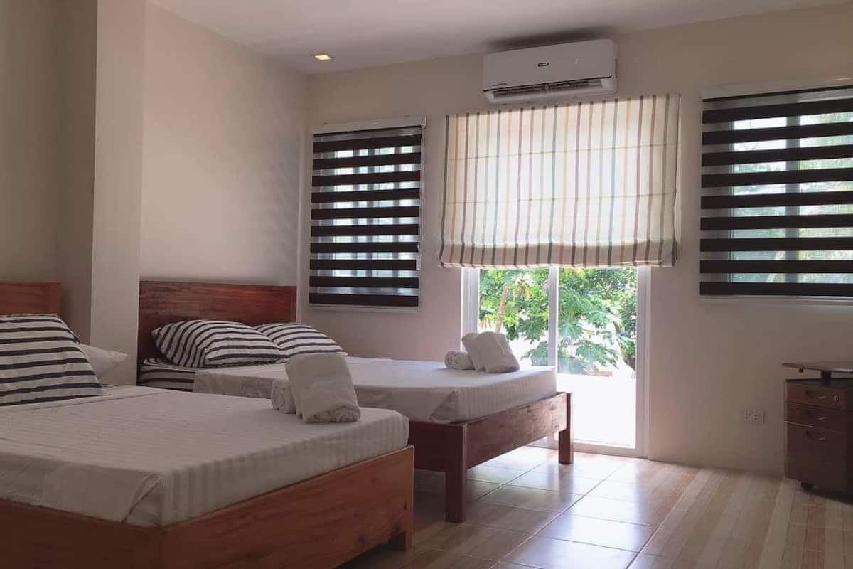 A bedroom with two beds, a wooden headboard, striped pillows, a window with blinds, and an air conditioning unit.