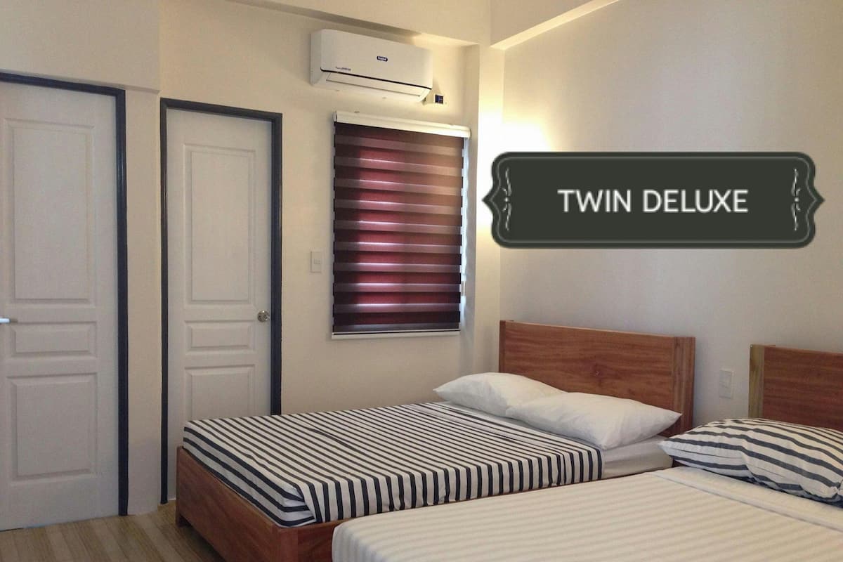 A twin deluxe room with two beds, striped bedding, a window with blinds, an air conditioning unit, and a sign indicating the room type.