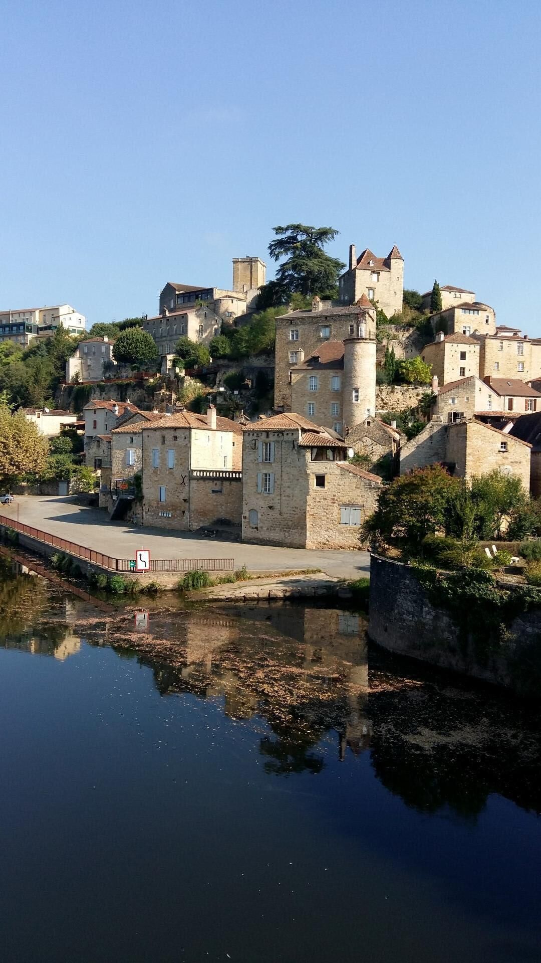 Une ville historique aux bâtiments en pierre, un château perché sur une colline et une rivière paisible en premier plan.