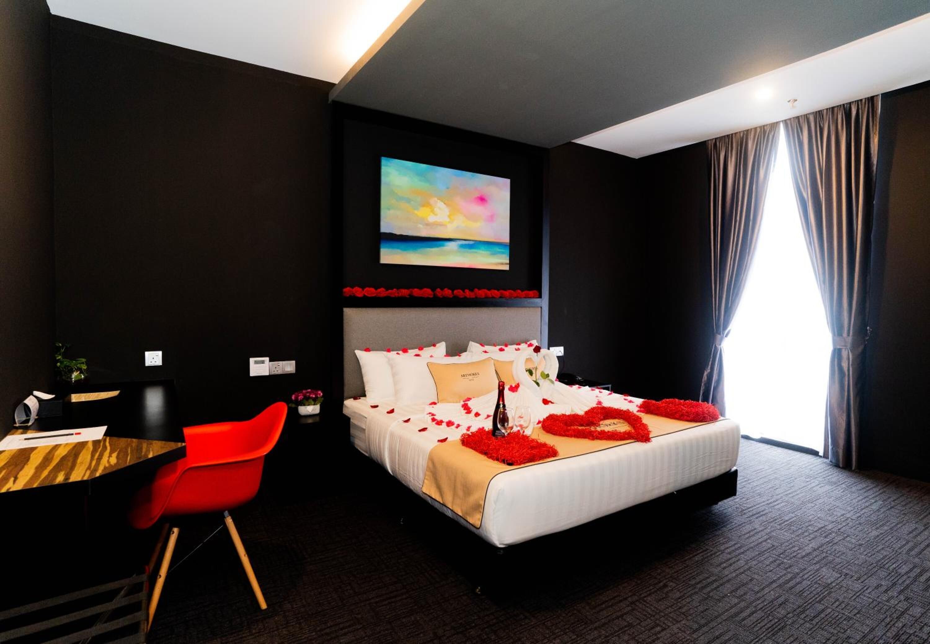A hotel room with a large bed, a desk with a chair, a painting on the wall, and a window with curtains.