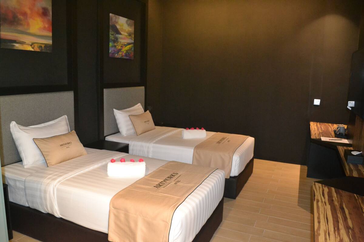 Two single beds with white linens and beige headboards, a wooden desk, and artwork on the walls.