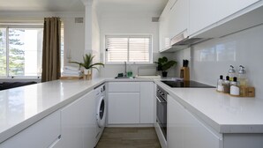 A modern kitchen with white cabinetry, a built-in washing machine, and a large window with blinds. - Manly Beachfront Apartment (Manly)