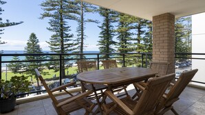 A balcony with a wooden table and chairs, overlooking a beach and forested area. - Manly Beachfront Apartment (Manly)