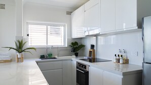 A modern kitchen with white cabinets, a stainless steel refrigerator, and a window with blinds. - Manly Beachfront Apartment (Manly)