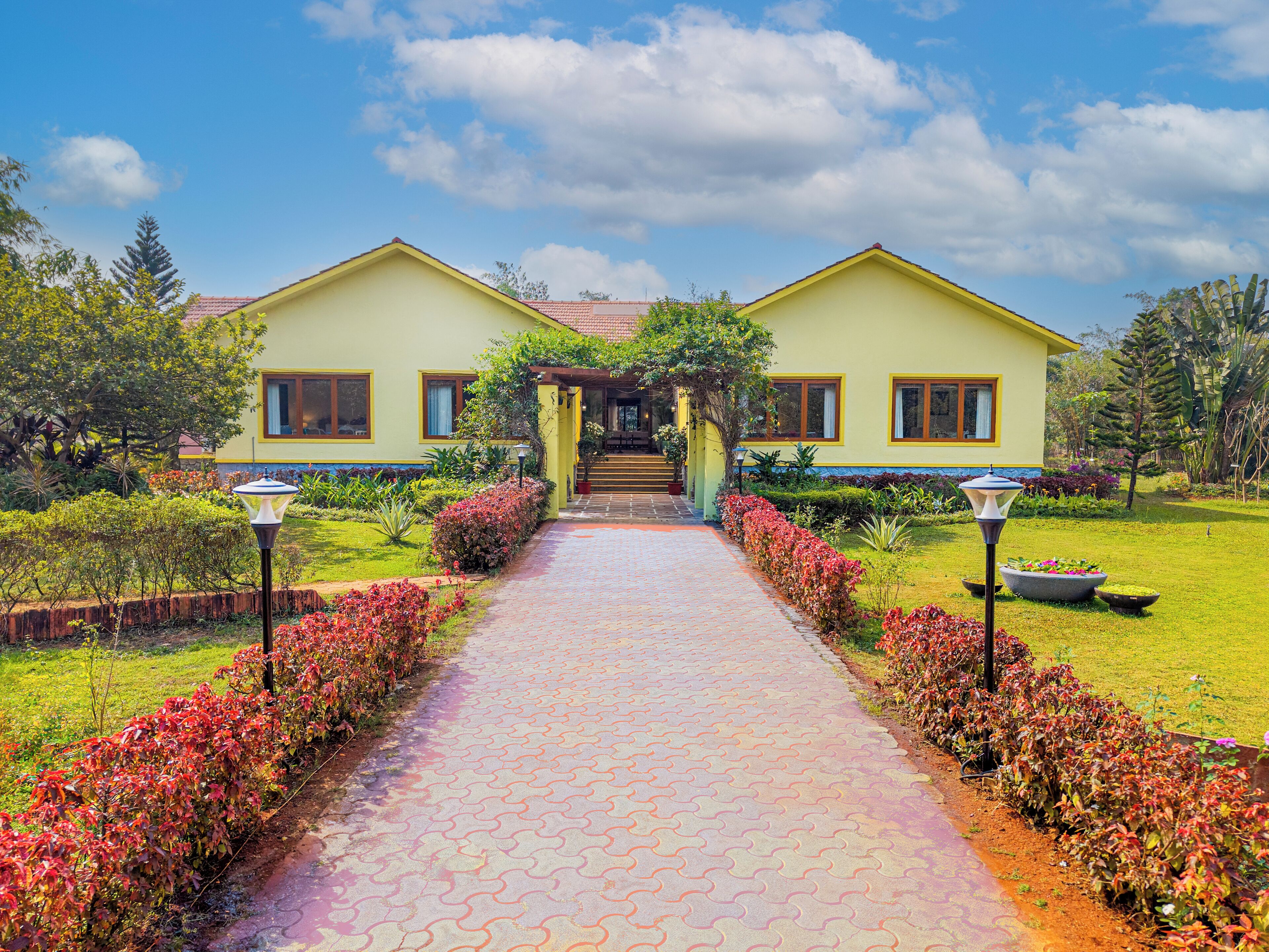 Edificio amarillo con un sendero empedrado que conduce a una entrada cubierta, rodeado de jardines y árboles bien cuidados.