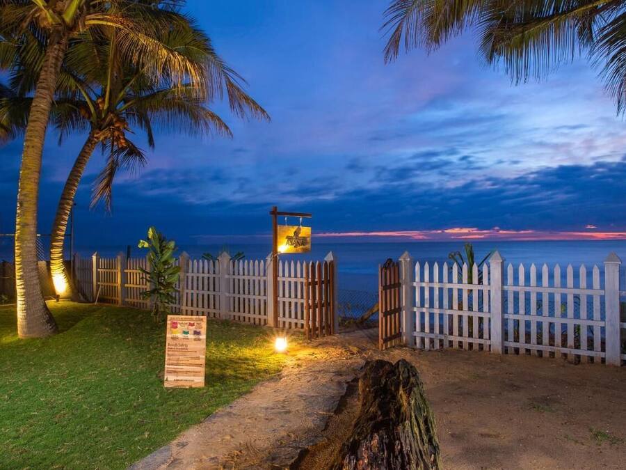 A beachside entrance with a wooden fence, palm trees, and a signboard.