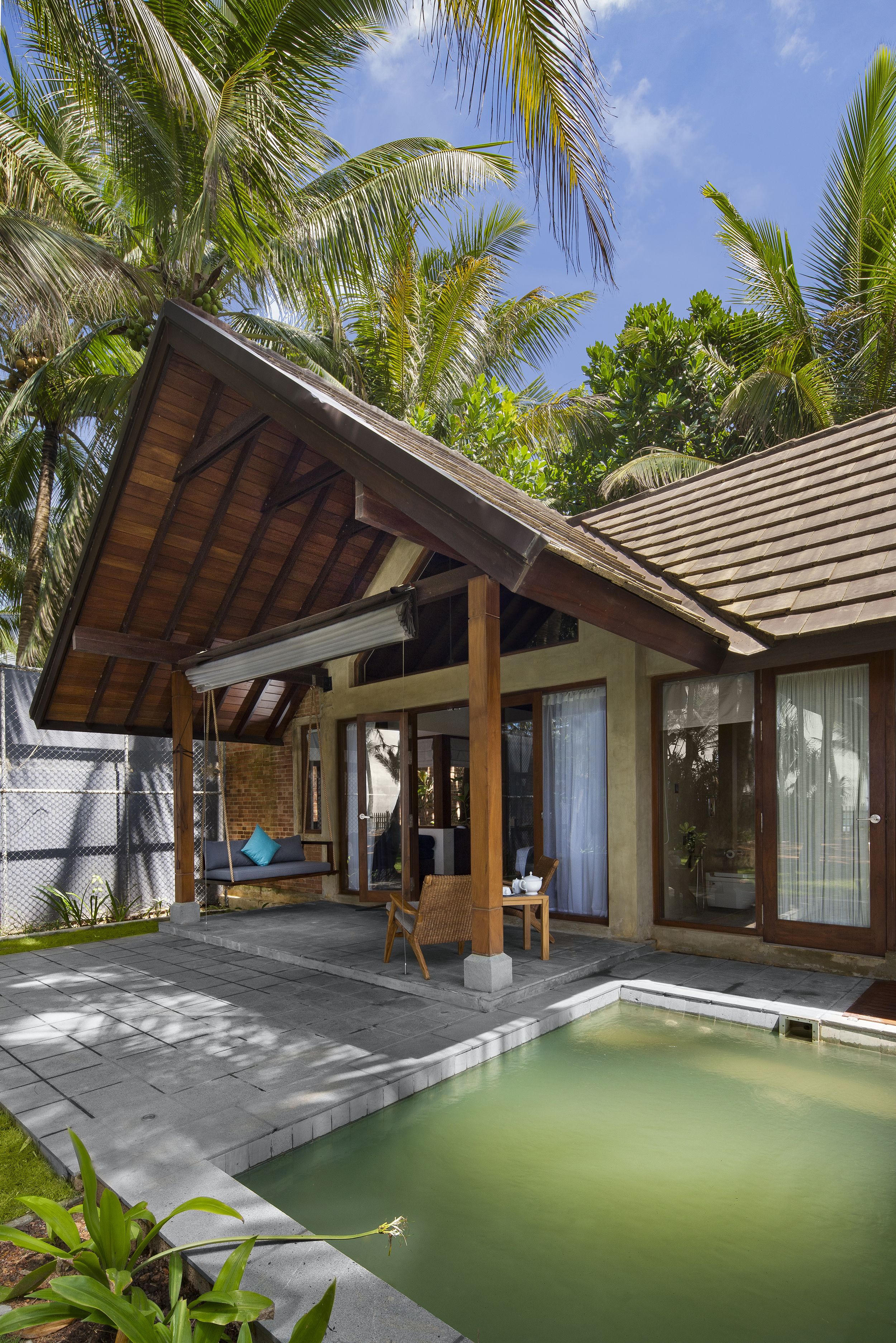 A covered outdoor seating area with a pool, palm trees, and a brick building.