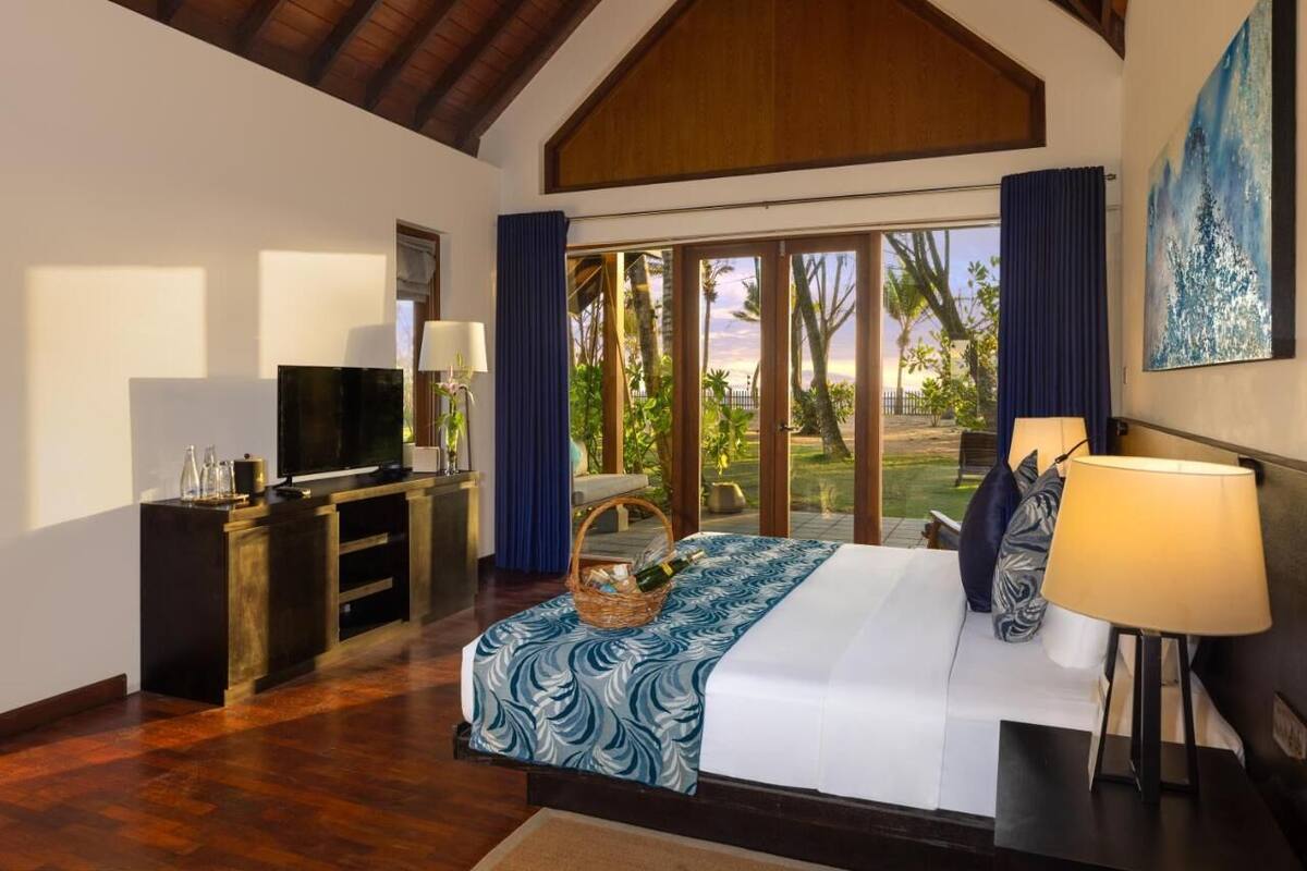 A bedroom with a large bed, a TV, a desk with a computer, and a view of the outdoors.