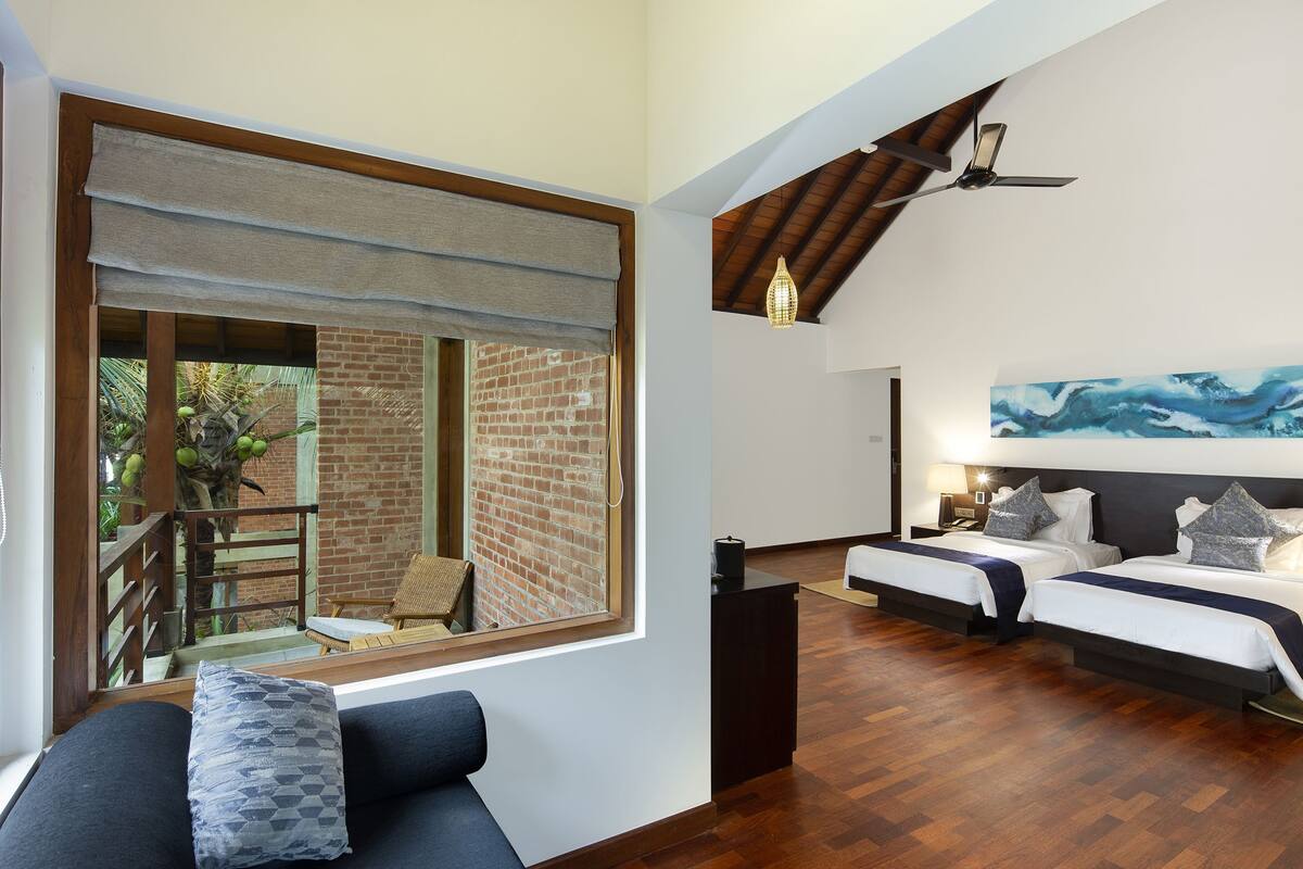 A bedroom with two beds, a large window, a ceiling fan, and a view of a brick wall and greenery outside.