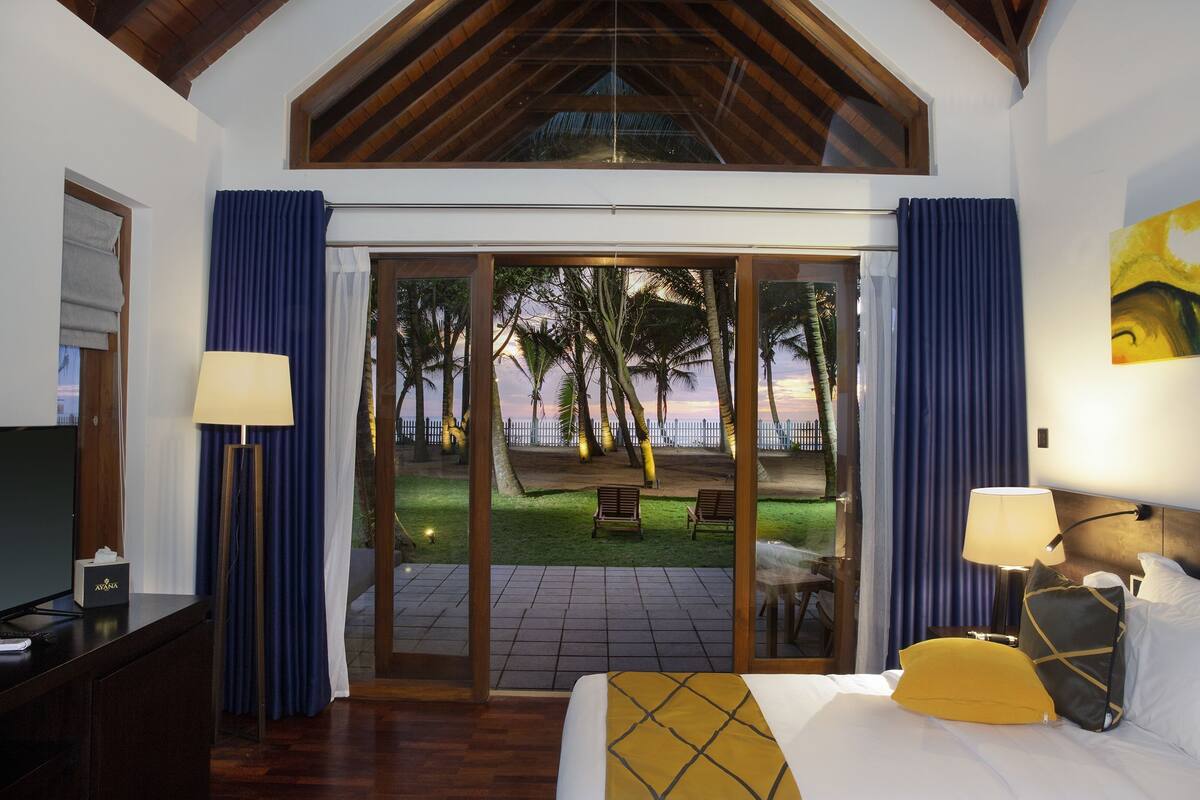 A hotel room with a bed, a TV, a desk, and a view of a palm tree garden through a glass door.