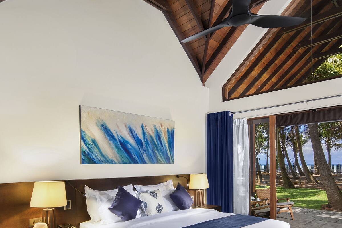 A bedroom with a bed, nightstands, a ceiling fan, a large abstract painting, and a view of palm trees through a window.