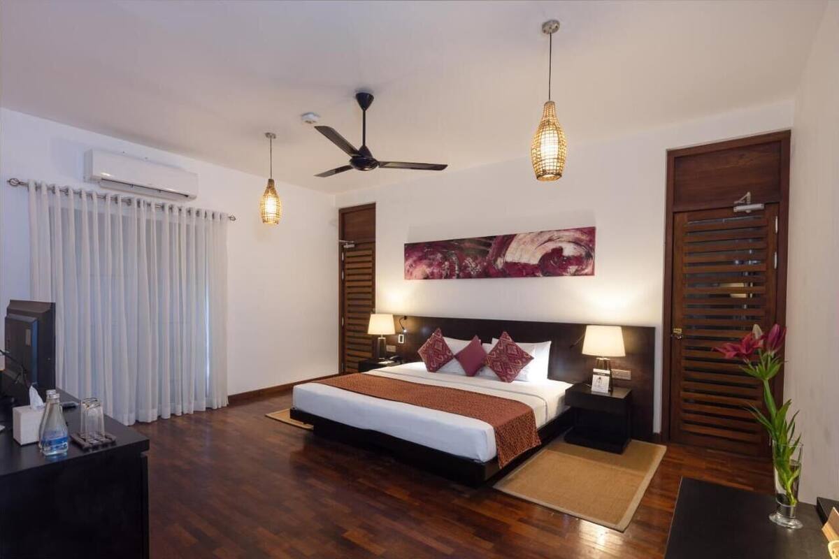 A hotel room with a large bed, ceiling fan, pendant lights, and a painting on the wall.