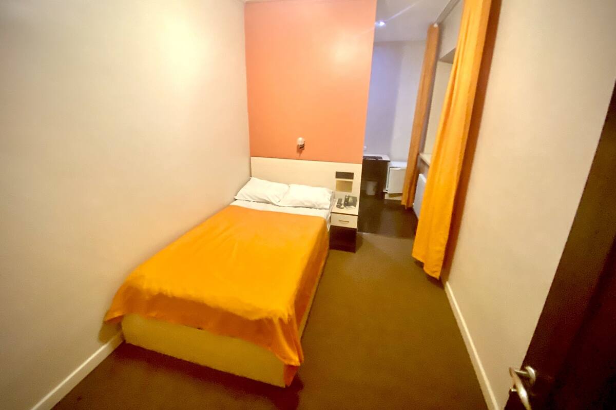 Une chambre d’hôtel étroite, avec un lit, un petit bureau et une table de chevet.