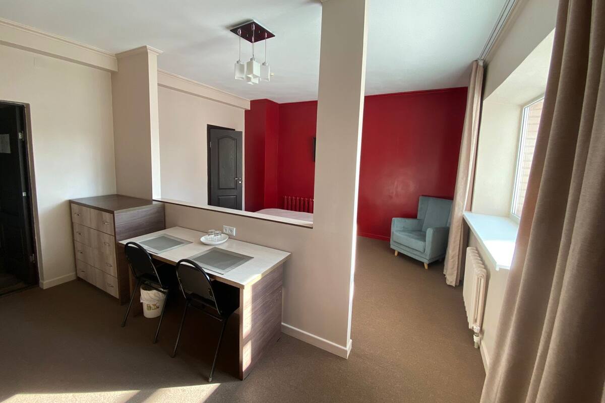 Une pièce compacte comprenant un coin cuisine, un bureau avec deux chaises, un mur d’accent rouge, une fenêtre avec des rideaux et un radiateur.