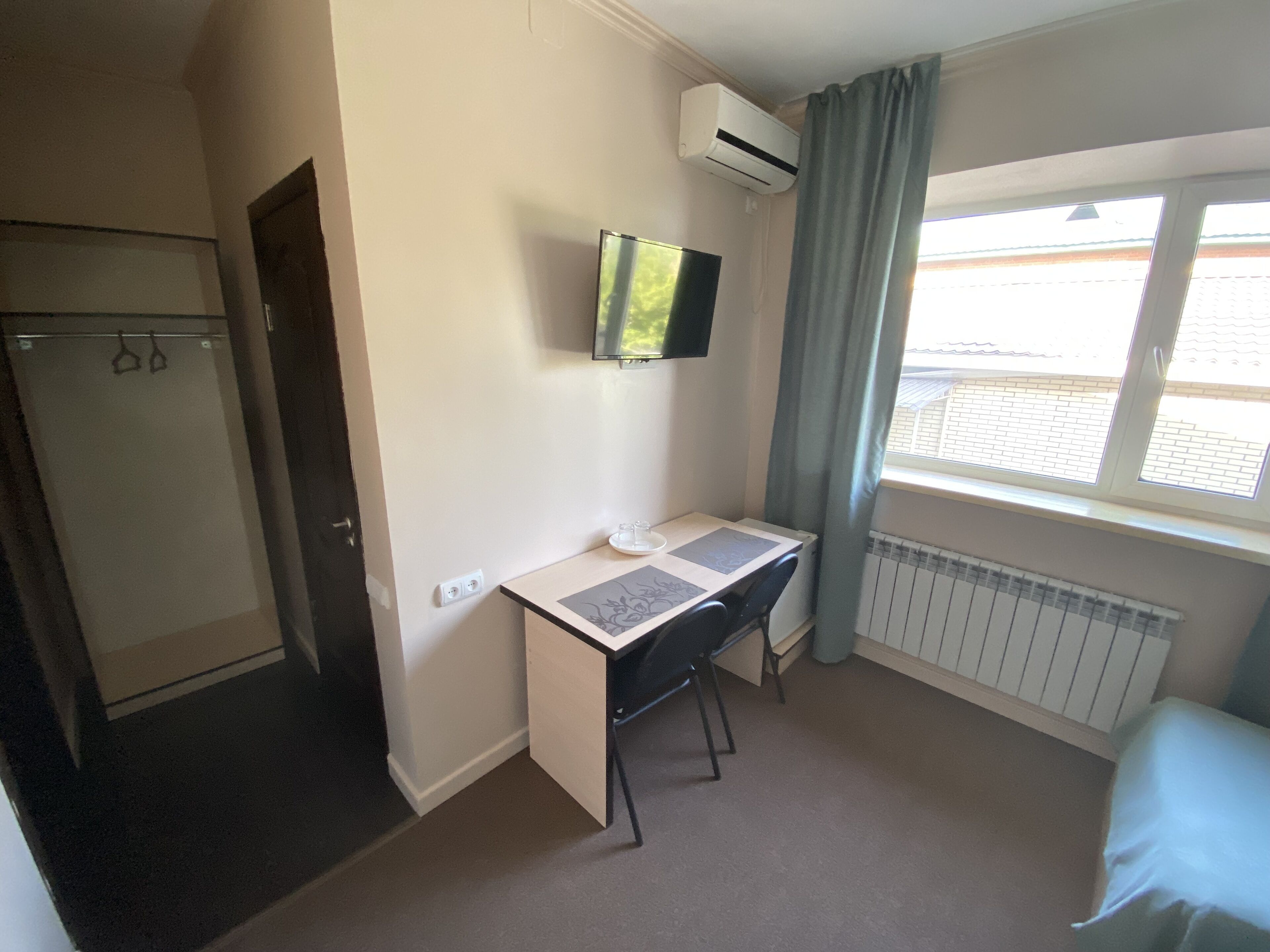 Une pièce de taille modeste, comprenant un bureau, un téléviseur fixé au mur, une fenêtre avec des rideaux, un radiateur et une porte.