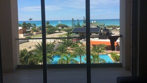 Blick aus einem Fenster auf einen Swimmingpool, einen Strand und ein Segelboot auf dem Wasser.