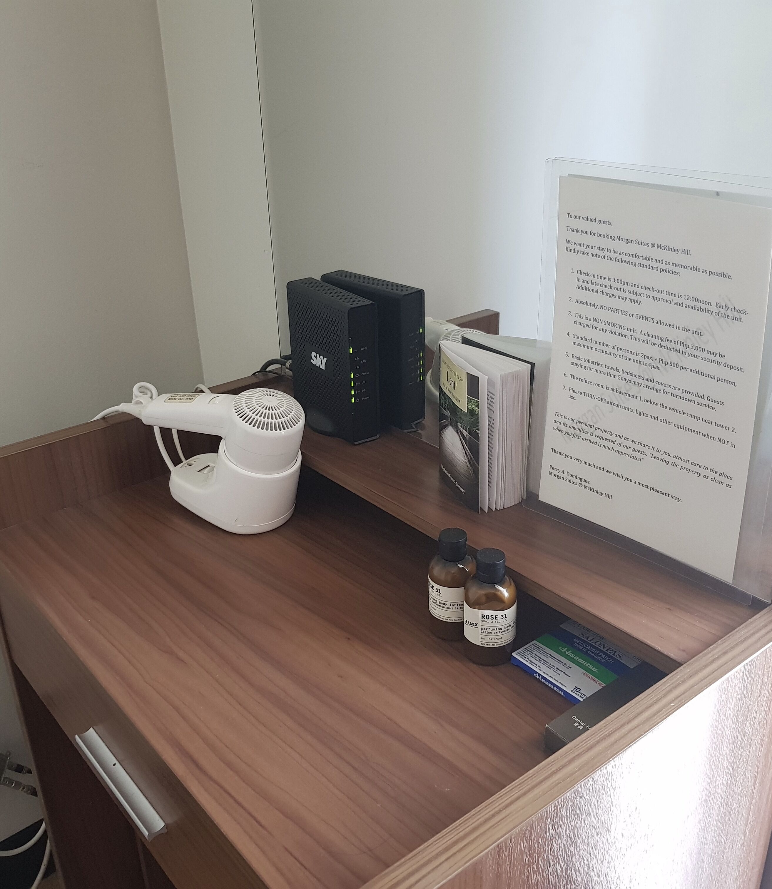 A hotel desk with a white hairdryer, two black Sky brand speakers, a stack of books, and two bottles labeled 'Rose 31'.