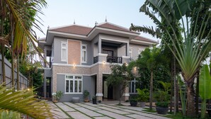 A modern two-story house with a balcony, surrounded by a well-maintained garden with various plants and a paved walkway.
