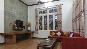 A living room with a wooden coffee table, a red sofa, a TV on a wooden stand, and a window with decorative curtains.