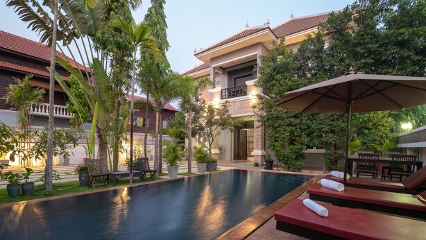 A swimming pool with lounge chairs and umbrellas, surrounded by potted plants and a building with a tiled roof.